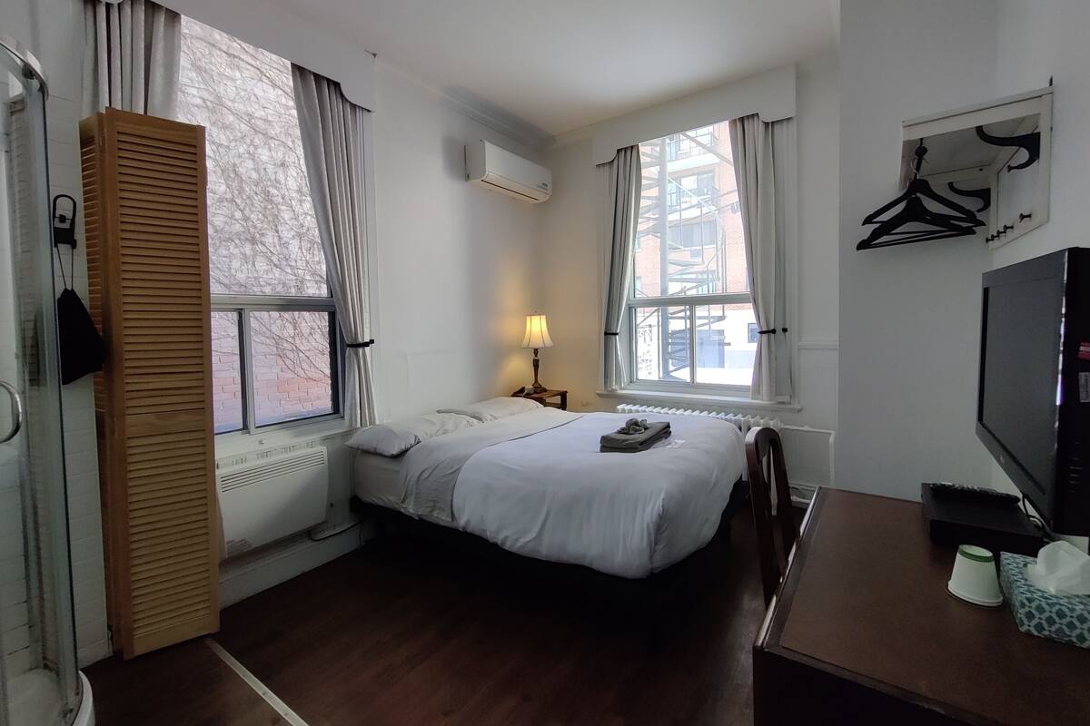 A hotel room with a bed, a desk, a TV, a radiator, and a window with curtains.