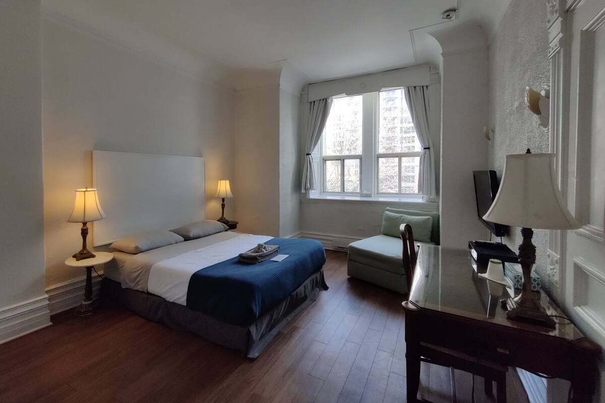 A bedroom with a bed, two bedside tables with lamps, a chair, a window with curtains, and a television on a stand.
