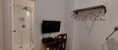 A room with a shower, a desk with a flat-screen TV, a chair, a wooden shelf with hangers, and a desk lamp.