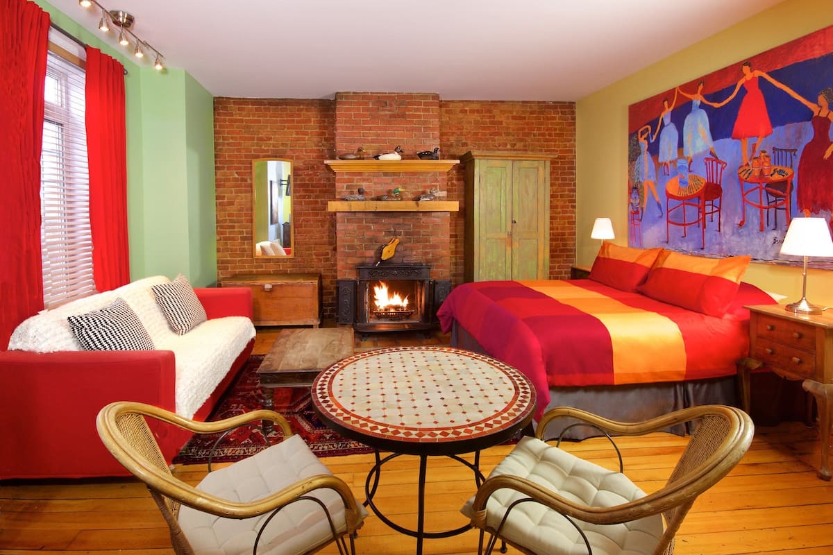 Una habitación acogedora con chimenea, una cama con ropa de cama roja, un sofá rojo, sillones de mimbre y un mural colorido en la pared.