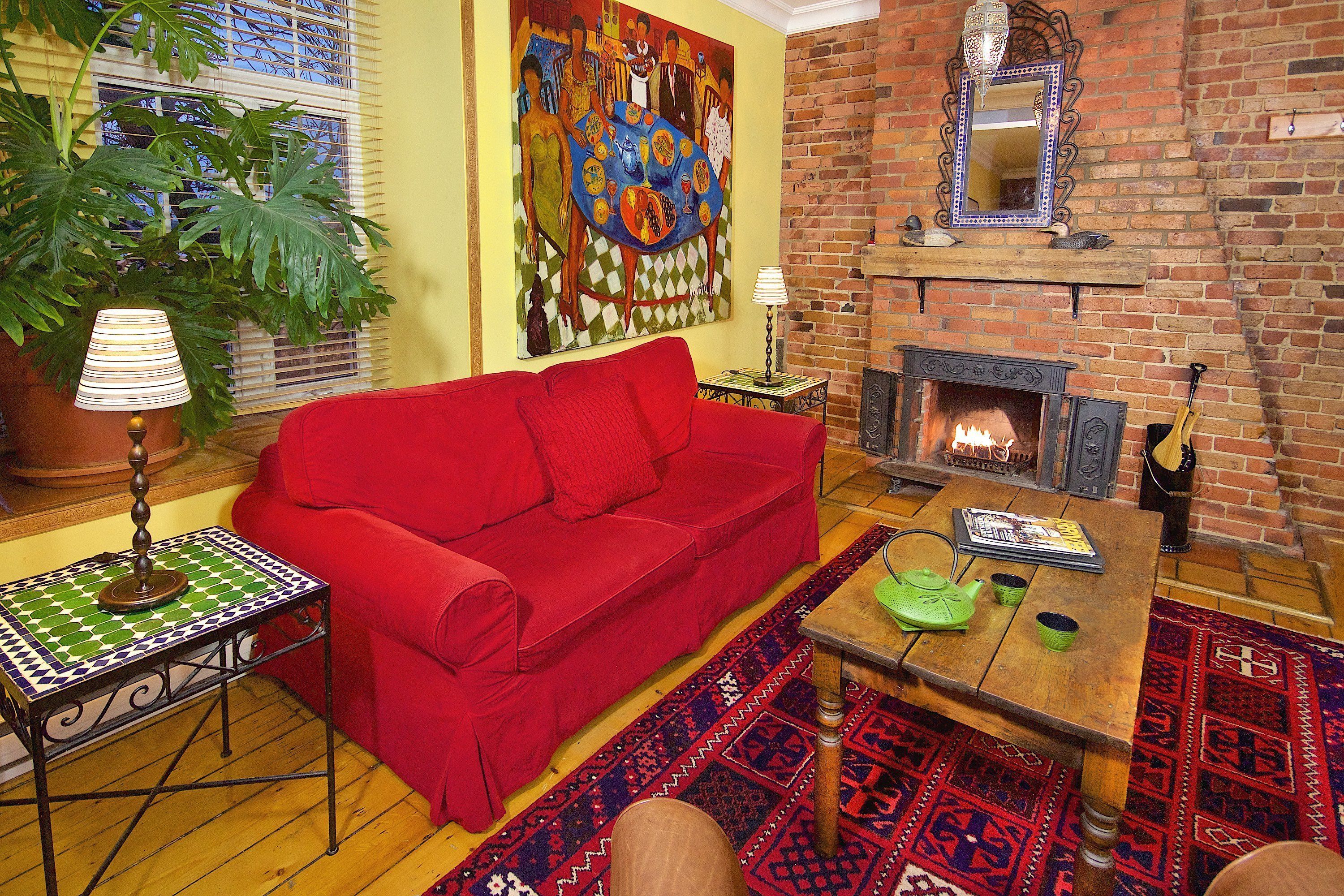 A cozy living room with a red sofa, a wooden coffee table, and a patterned rug. A large painting adorns the wall, and a brick fireplace is visible.