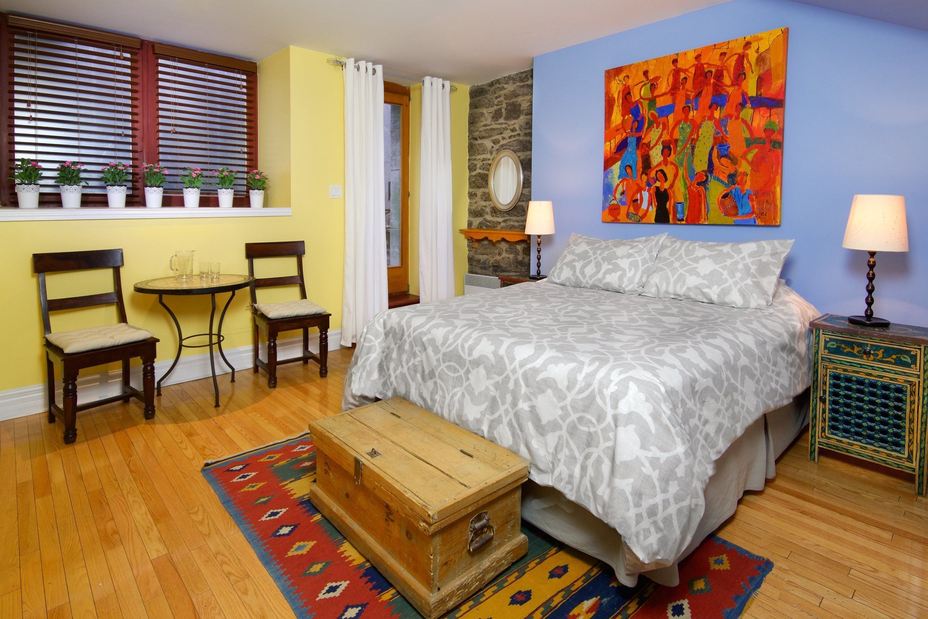 A bedroom with a bed, two chairs, a small table, a wooden chest, and a colorful rug.