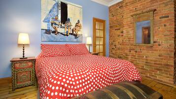 A bedroom with a red polka dot bedspread, a wooden bench, a painting on the wall, and a brick wall.