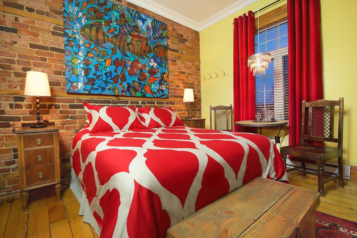 Una habitación con pared de ladrillo, un cuadro grande de un pez colorido, una cama con colcha de rayas rojas y blancas, una mesita de noche de madera, una mesa de madera y una silla.
