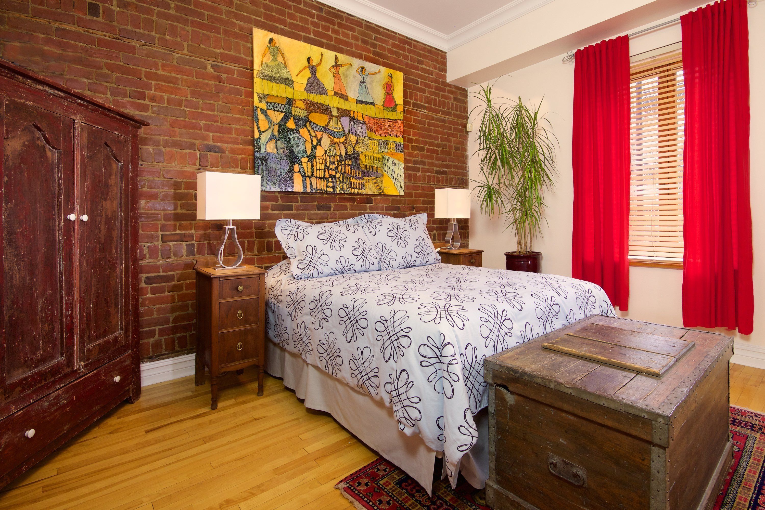 Une chambre avec un mur en briques, un lit recouvert d’une couvre-lit à motifs, une table de chevet en bois, une commode, un rideau rouge et une fenêtre avec des stores.