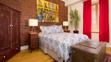 Une chambre avec un mur en briques, un lit recouvert d’une couvre-lit à motifs, une table de chevet en bois, une commode, un rideau rouge et une fenêtre avec des stores.