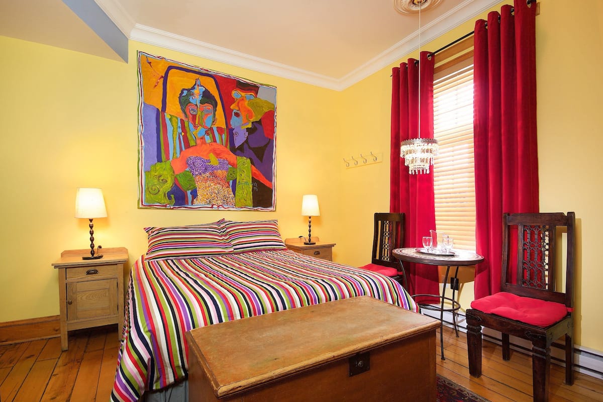 Habitación de hotel con una cama grande, dos mesitas de noche con lámparas, un banco de madera, una silla, una cortina roja y un cuadro abstracto colorido en la pared.