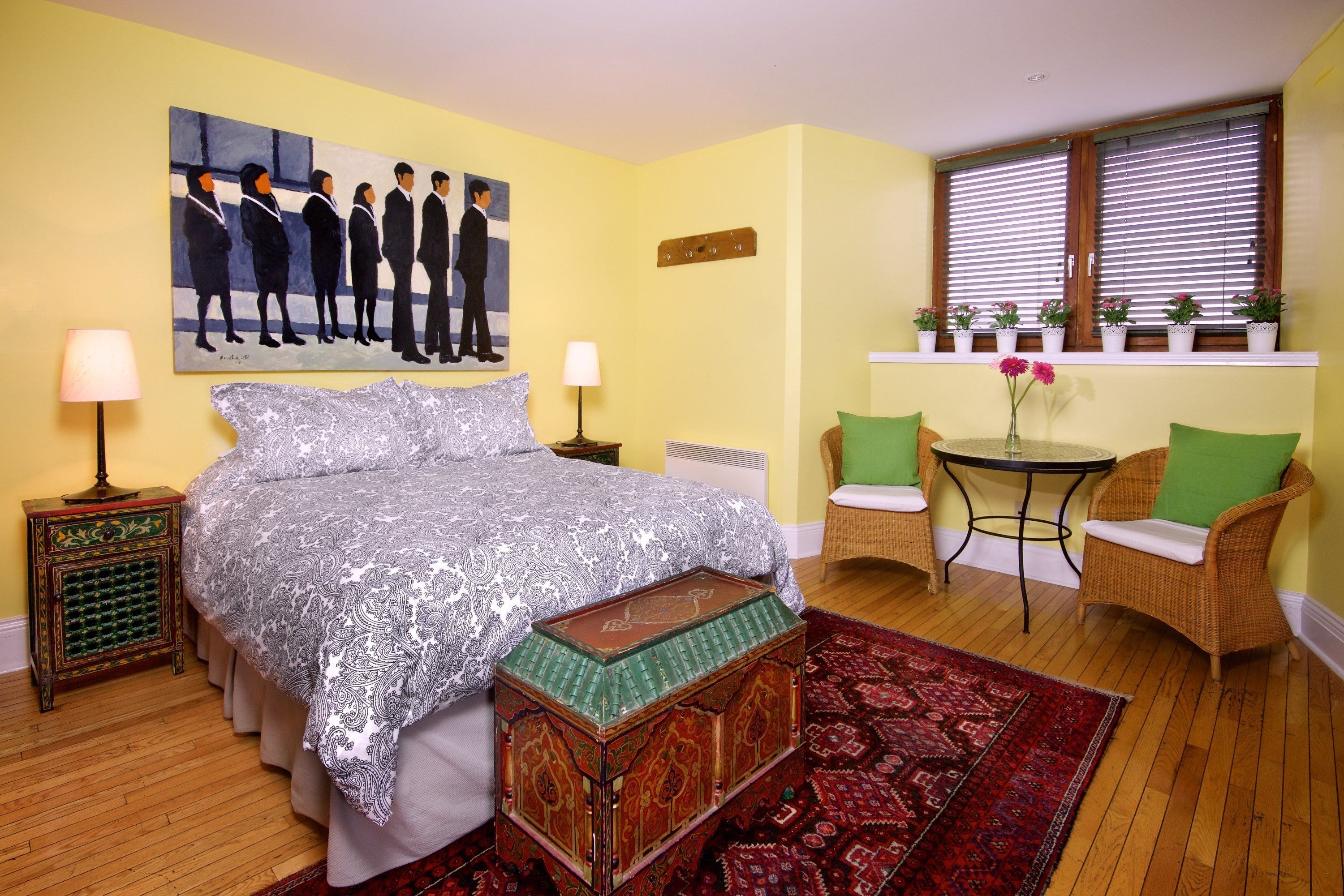 Un dormitorio con cama, mesitas de noche, una silla, una pequeña mesa y una ventana con persianas.