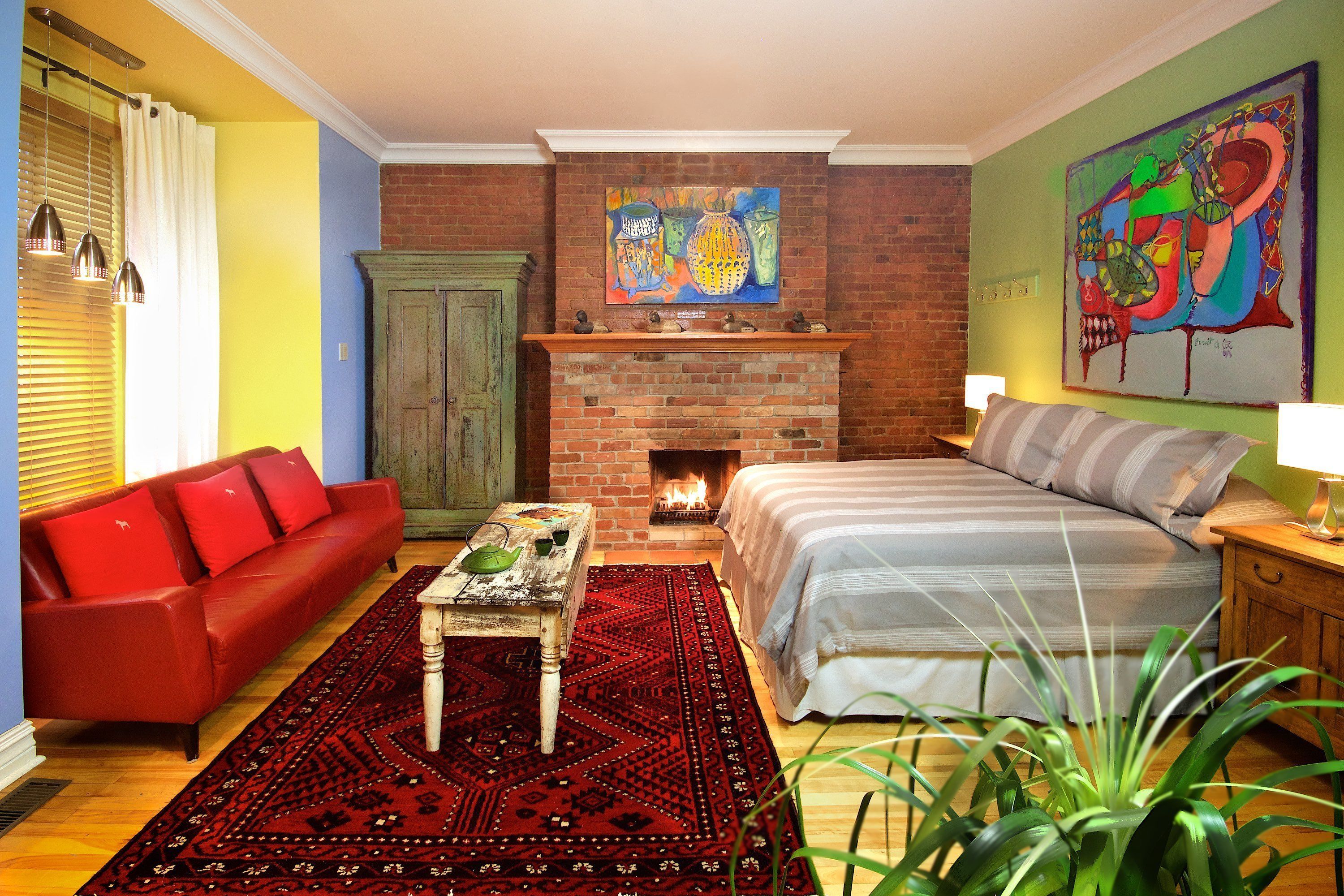 Uma sala de estar aconchegante com lareira de tijolos, sofá vermelho, uma cama grande e um quadro abstrato colorido na parede.