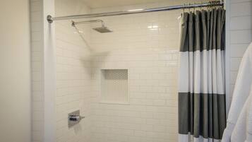 Ducha con azulejos blancos, cabezal de ducha tipo lluvia y cortina de baño a rayas grises y blancas.