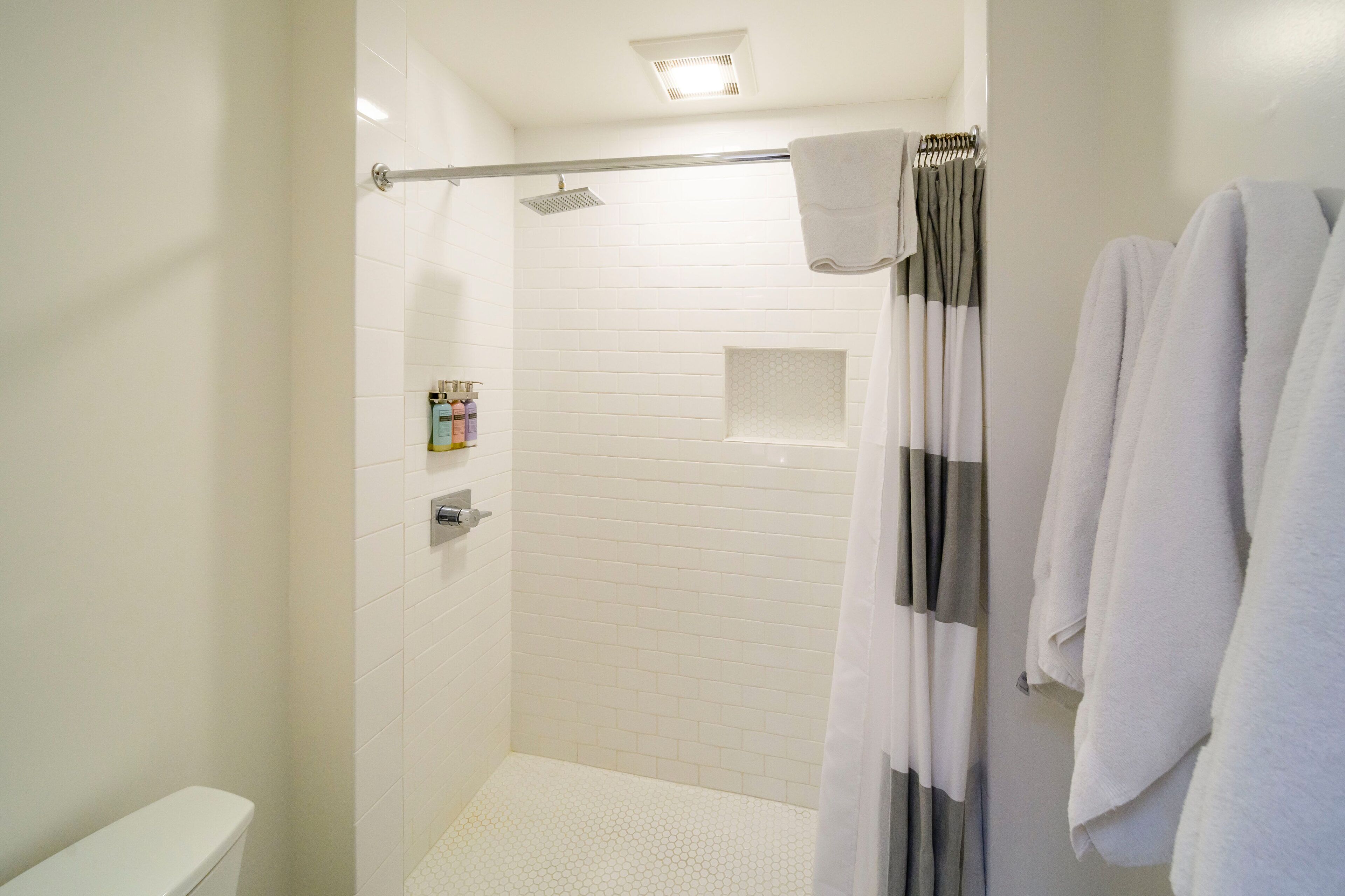 Un baño moderno con paredes de azulejos blancos, zona de ducha, inodoro y un perchero con toallas.