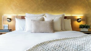 A neatly made bed with white linens and multiple pillows, a bedside table with a clock and books, and a wall-mounted lamp.