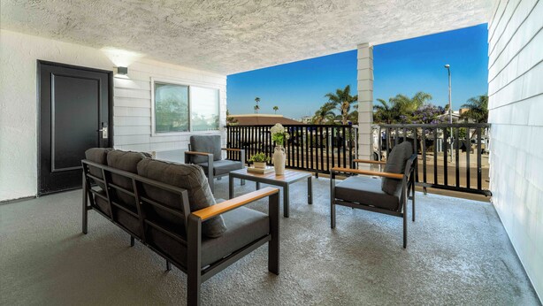A balcony with a sofa, chairs, and a small table, overlooking a clear blue sky and palm trees.