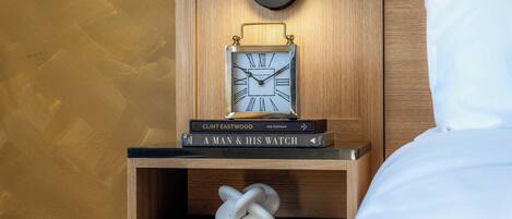 Una mesita de noche de madera con un reloj fijado a la pared, algunos libros y un objeto decorativo.