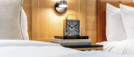 Una cama bien tendida con ropa blanca, un cabecero de madera, una mesita de noche con un reloj y una lámpara.