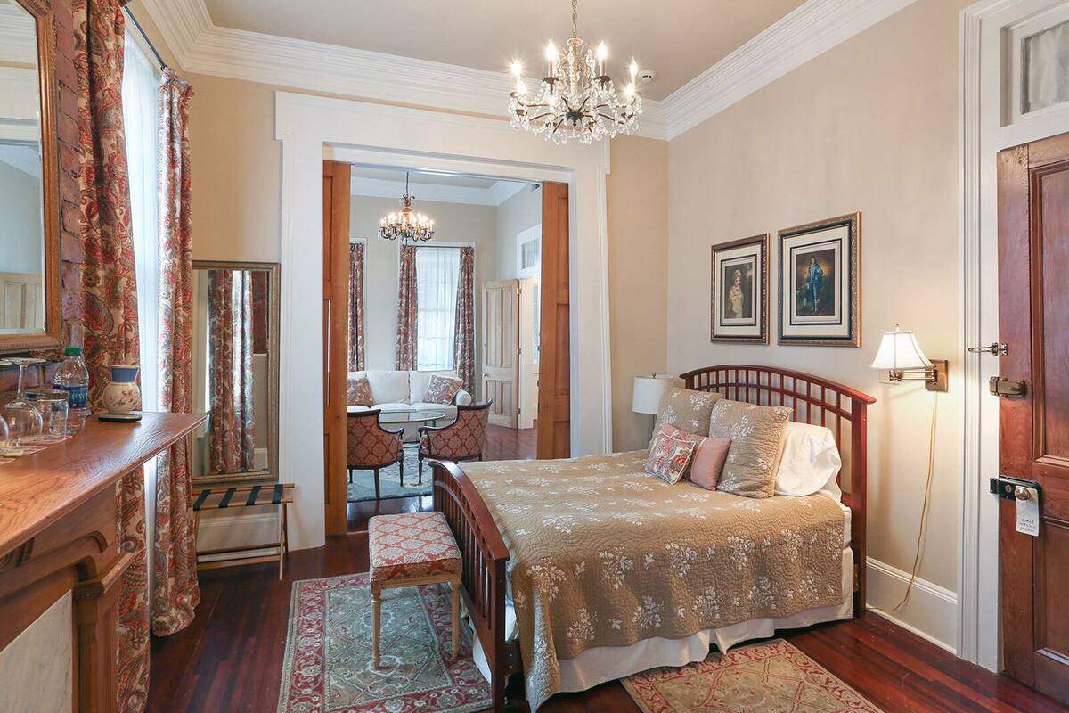 Une chambre à coucher avec un grand lit, une tête de lit en bois, un lustre, une table de chevet, un miroir et une porte.
