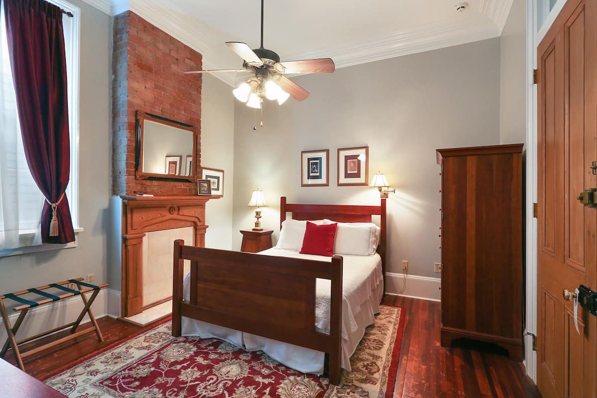 Une chambre avec un lit en bois, un ventilateur de plafond, un mur en briques, des tableaux encadrés et une porte en bois.