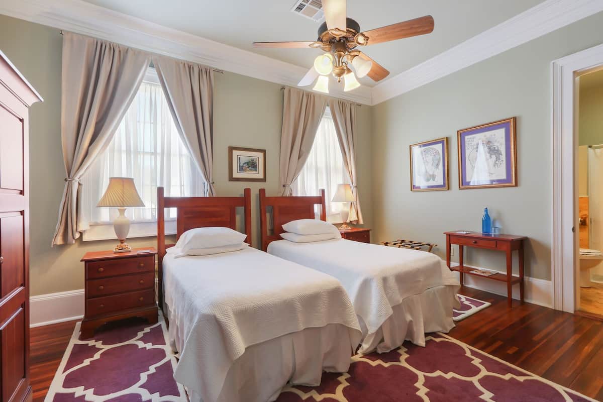 Une chambre d’hôtel avec deux lits, un ventilateur de plafond, une commode en bois, une table de chevet, une petite table et une salle de bain visible à travers une porte ouverte.