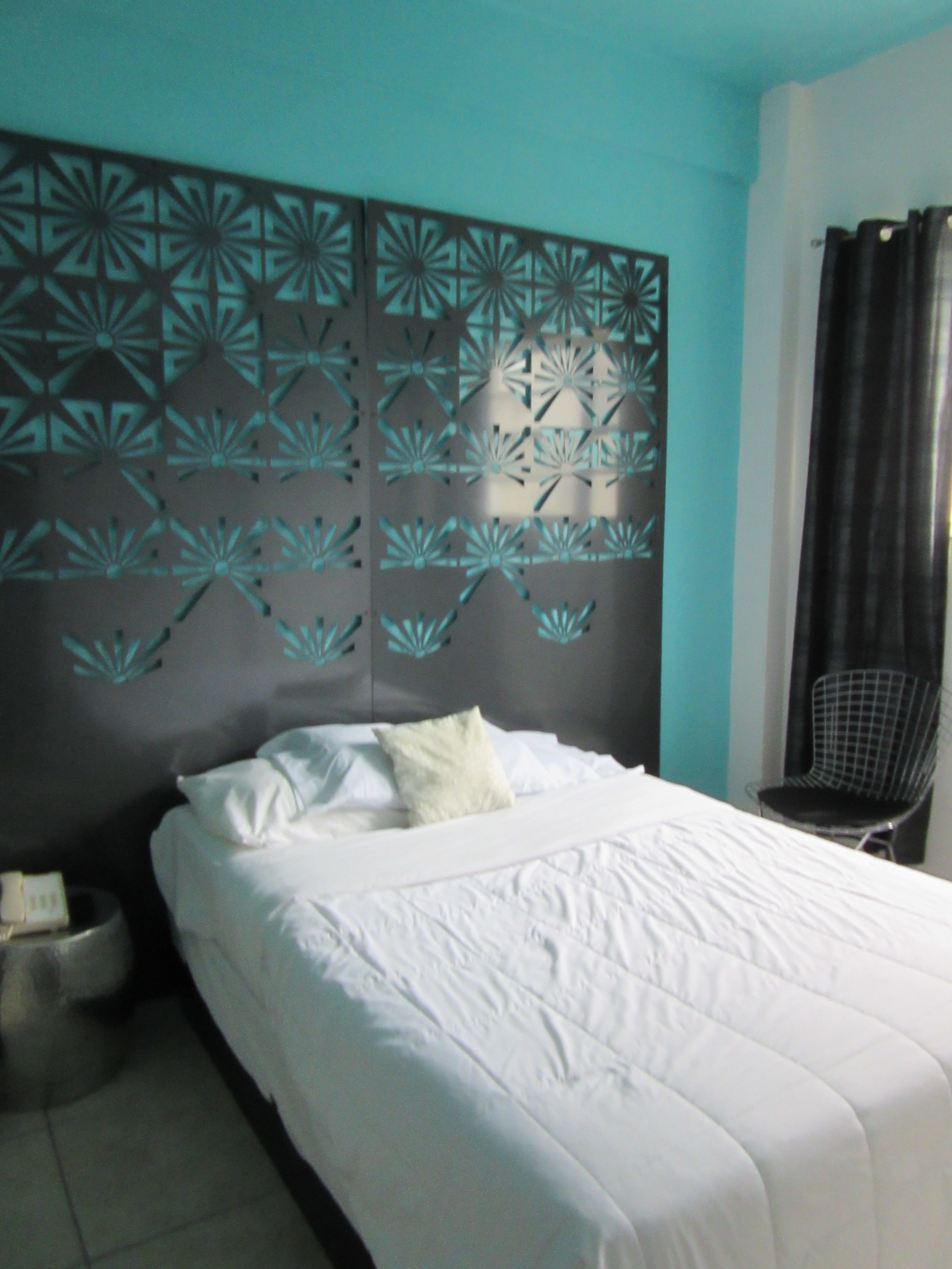 Una cama bien hecha con sábanas blancas, un cabecero decorativo con diseños geométricos, una silla y una mesita de noche con una lámpara.