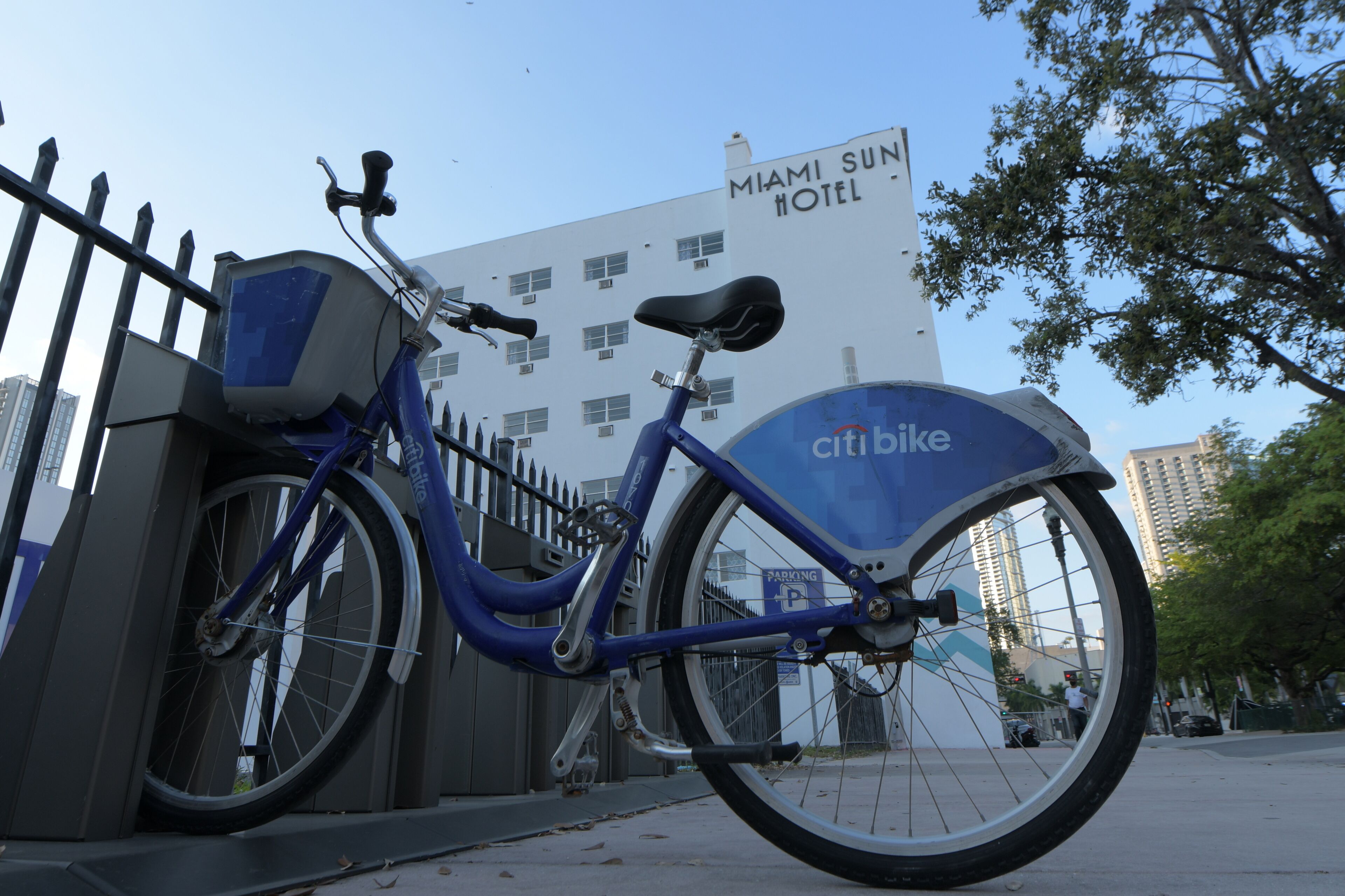 A CitiBike parked near a fence with a Miami Sun Hotel in the background.