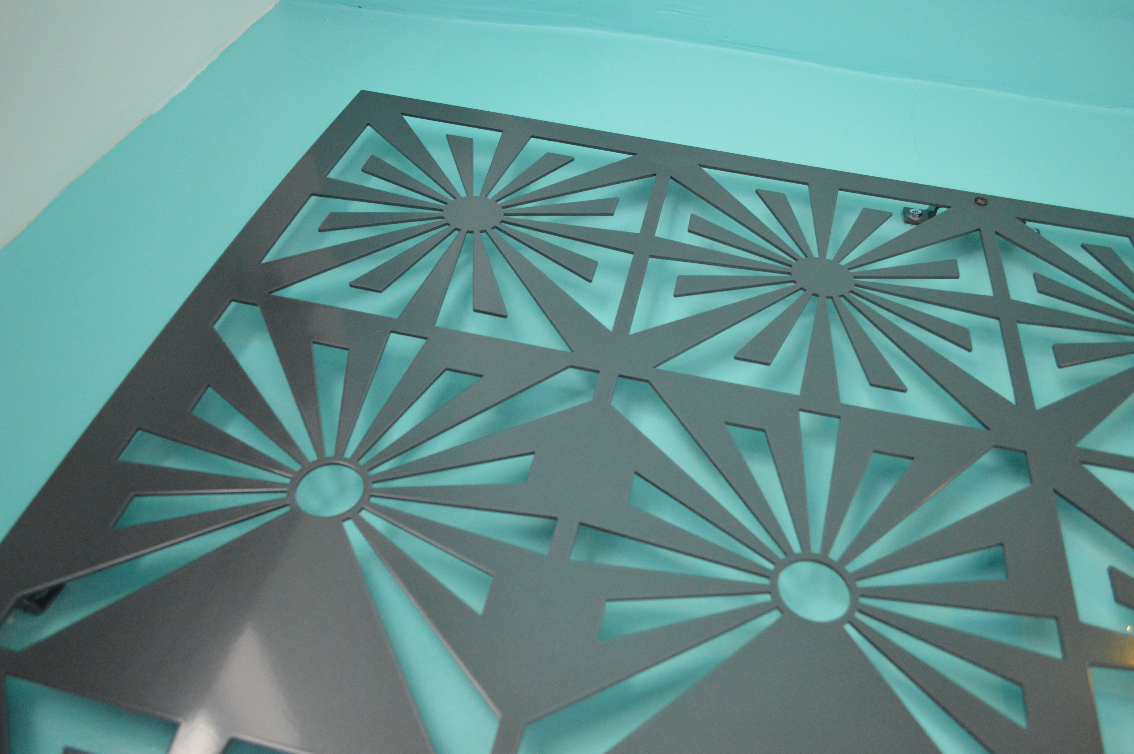 A geometric metal panel with a central circular design and radiating lines, mounted on a teal background.