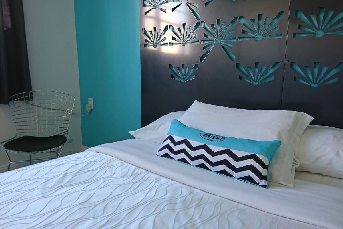 A bed with white bedding and a patterned pillow, a metal chair, and a wall with decorative cutouts.