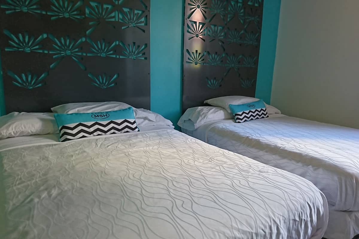 Two beds with white bedding and teal pillows, a teal accent wall, and a decorative panel with a geometric pattern.