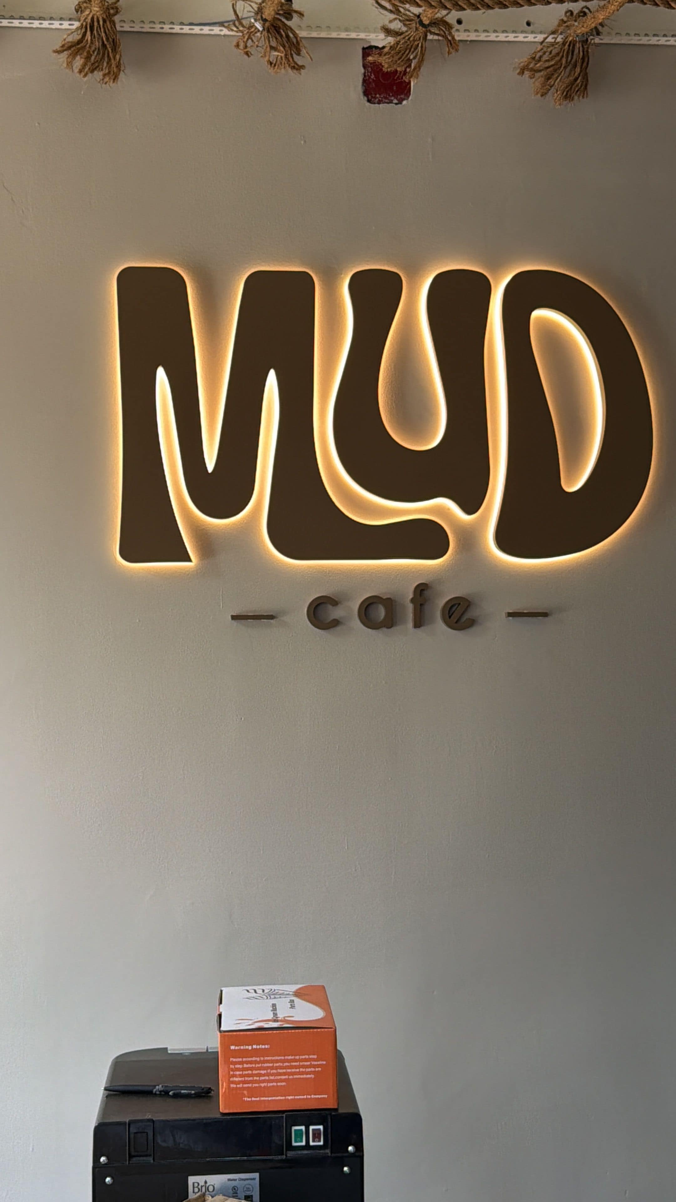 Een muur met verlichte letters die 'MUD cafe' spellen en een printer met een doos erop.