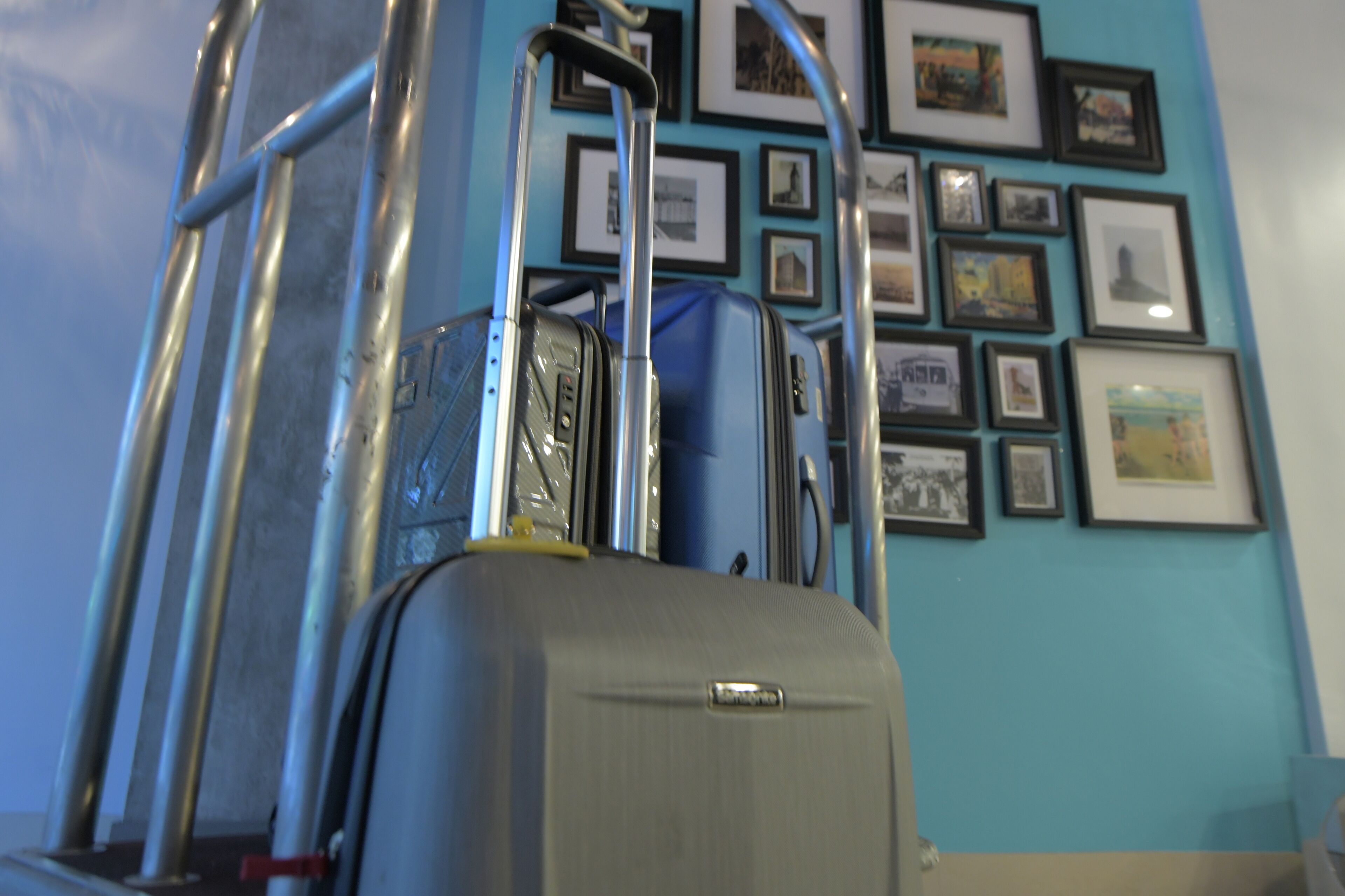 A room with a wall decorated with framed pictures, a luggage cart, and a suitcase.