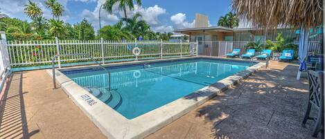 A swimming pool with a white fence, lounge chairs, and palm trees in the background.