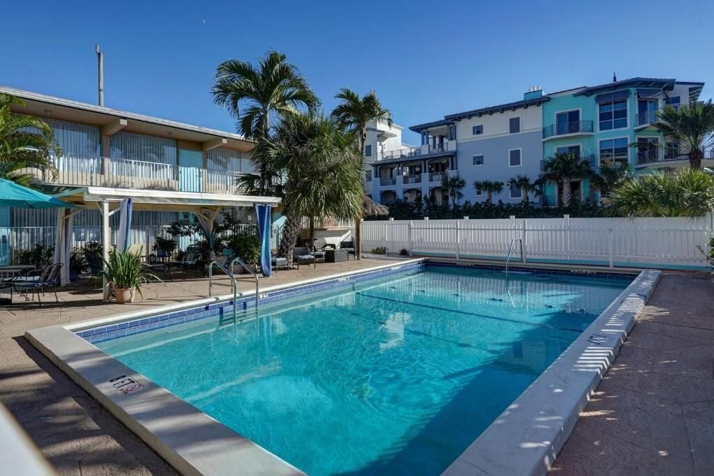 A swimming pool with a white fence, palm trees, and a covered seating area with umbrellas.
