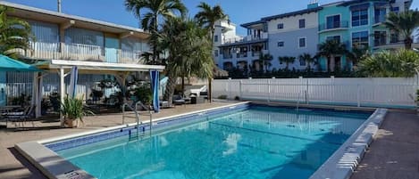 A swimming pool with a white fence, palm trees, and a covered seating area with umbrellas.