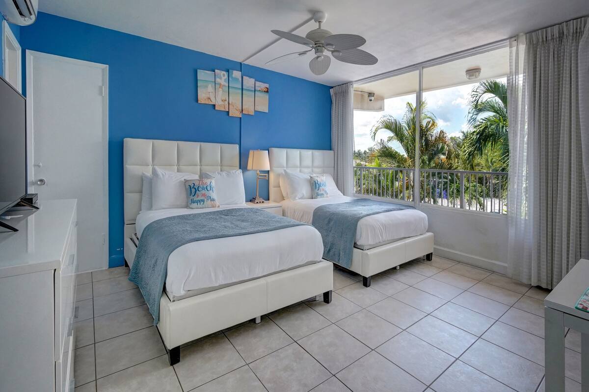 A hotel room with two beds, a ceiling fan, a balcony with a view of palm trees, and a large window.