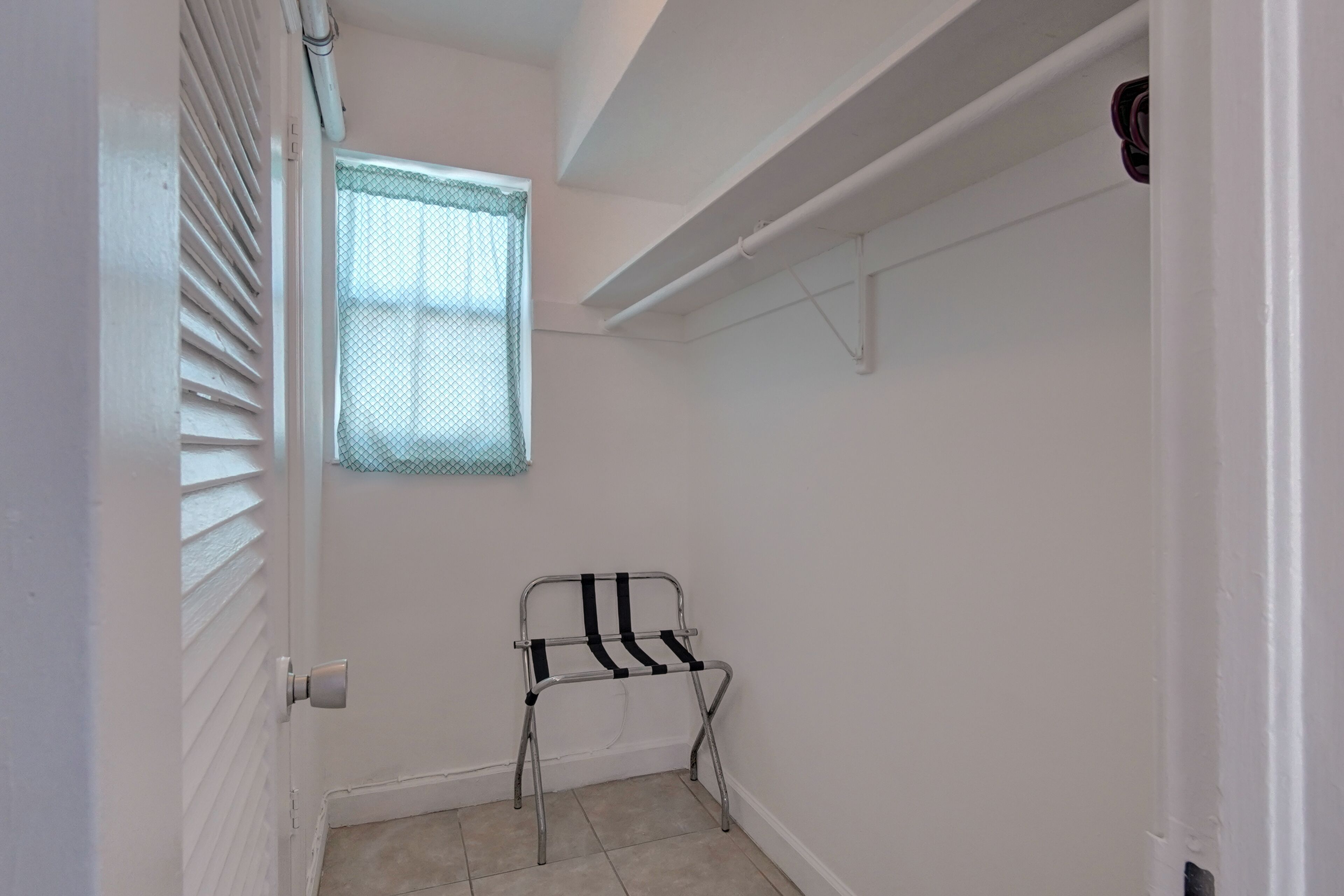 A narrow hallway with white walls, a window with a mesh screen, a metal chair, and a white door.