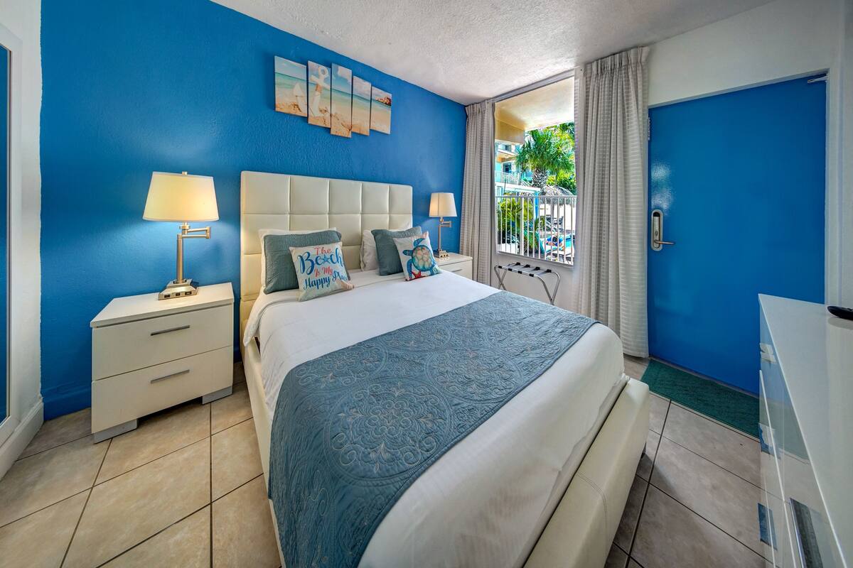 A hotel room with a large bed, two bedside tables, a nightstand, a window with curtains, and a view of a pool and greenery outside.