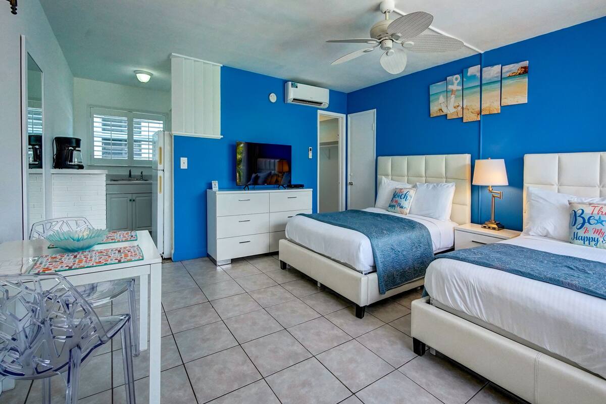 A hotel room with two beds, a dining area with a table and chairs, a kitchen area, and a ceiling fan.