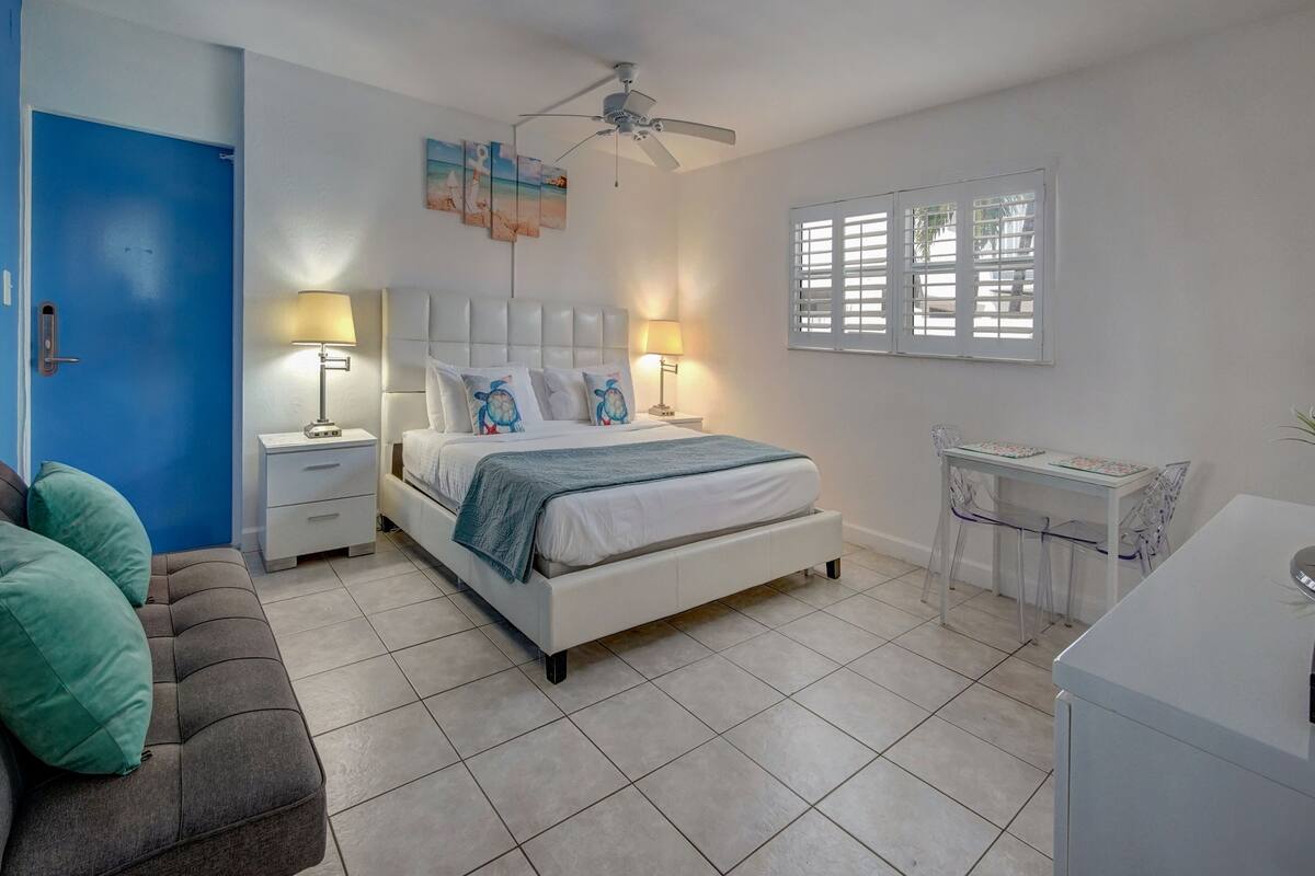 A bedroom with a bed, bedside tables, a desk, a chair, a ceiling fan, and a window with blinds.