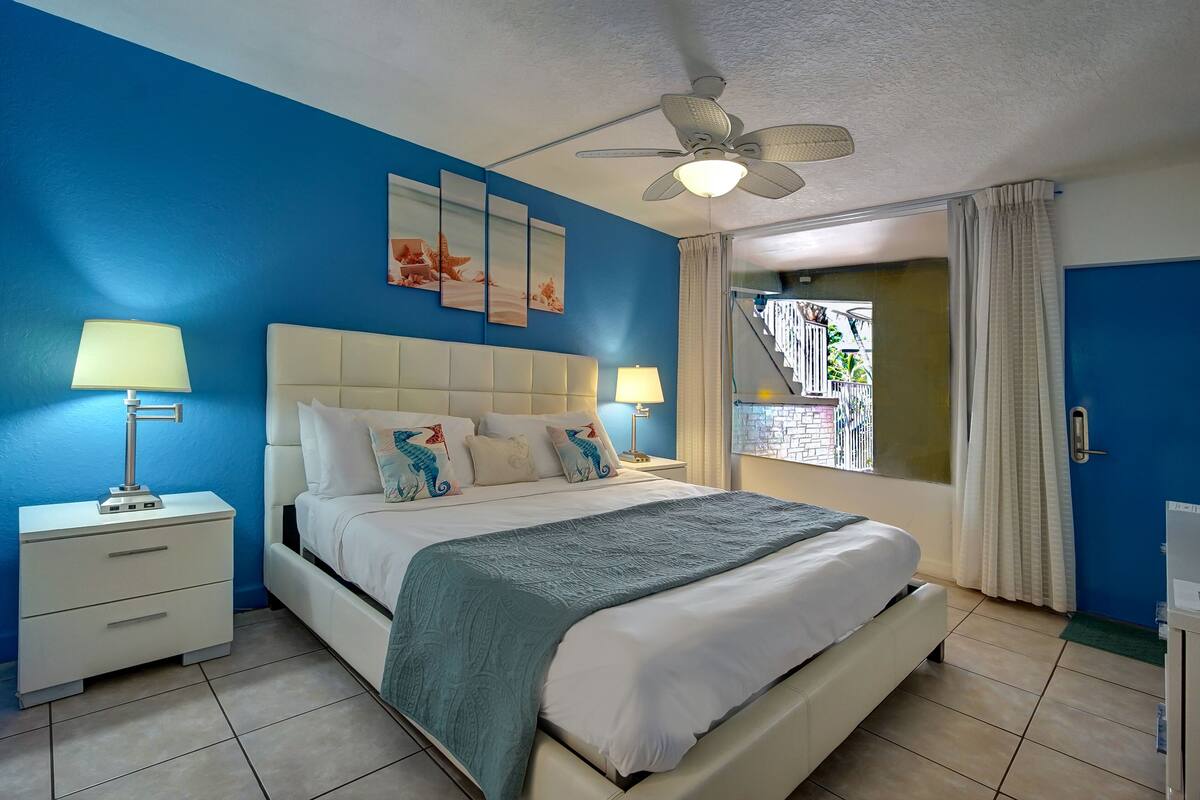 A bedroom with a bed, two bedside tables with lamps, a ceiling fan, and a window with curtains.