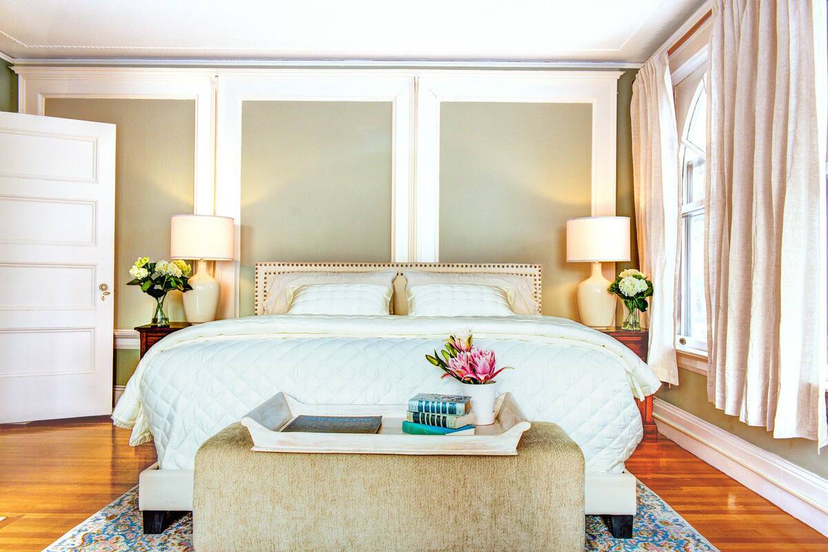 A neatly arranged bedroom with a large bed, bedside tables with lamps, a tray with books and flowers, and a door.