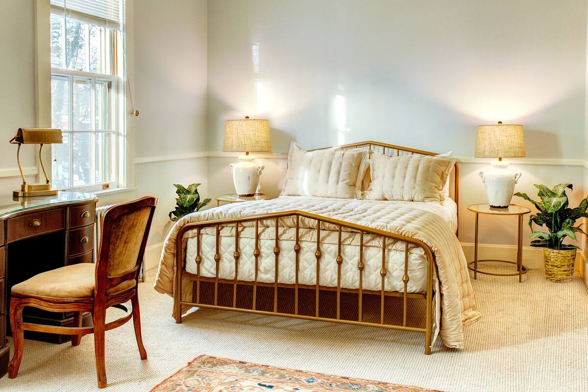 A bedroom with a bed, bedside lamps, a desk, a chair, and a window.