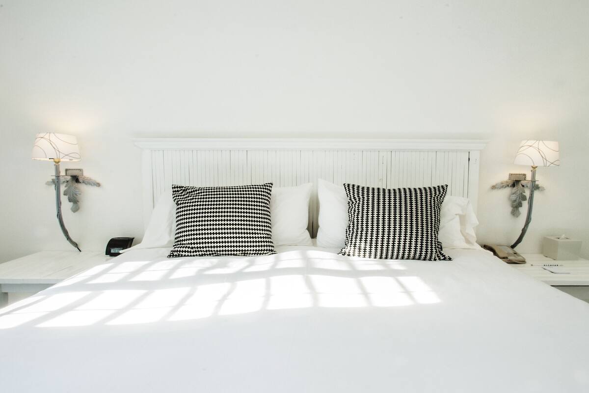 Una cama bien hecha con cabecera blanca, dos almohadas con estampado blanco y negro, y dos mesitas de noche con lámparas.