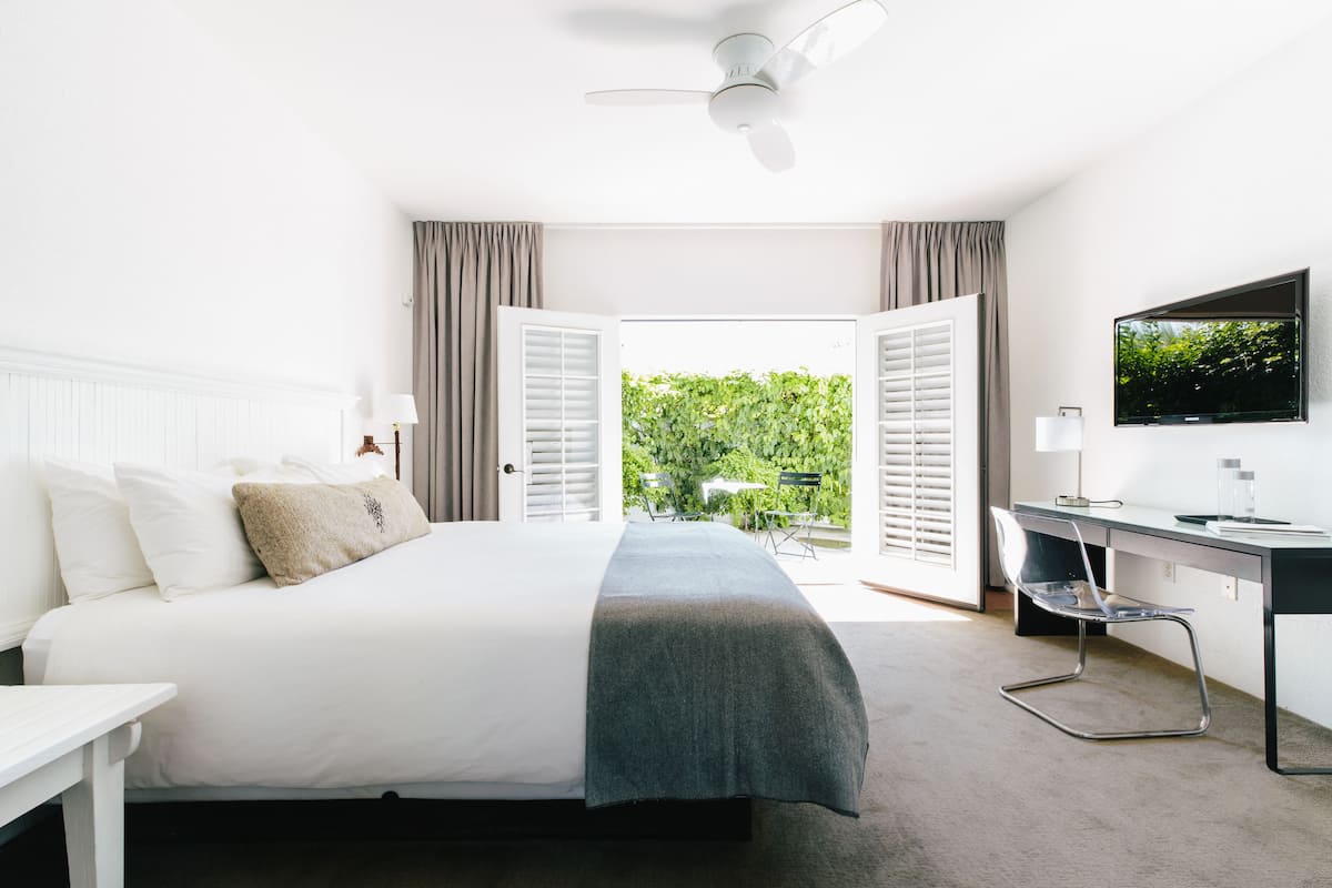 Un dormitorio con cama, escritorio, silla, televisor y vistas al exterior con vegetación.
