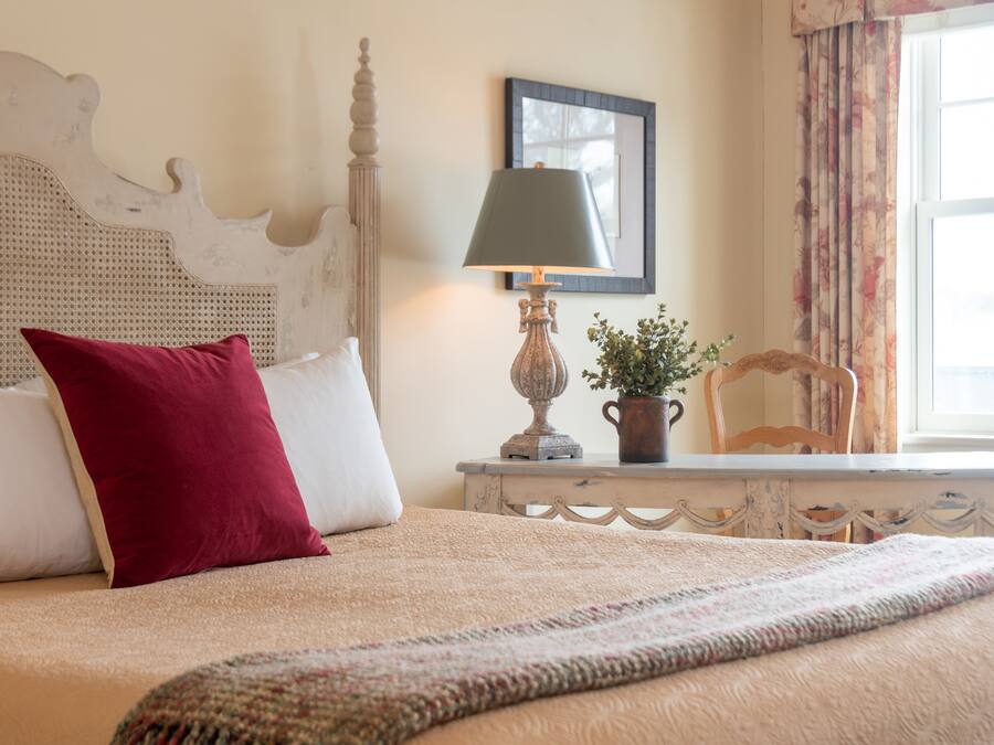 Una cama bien hecha con un almohadón rojo, un almohadón blanco y una manta color beige. Hay una lámpara con pantalla verde sobre una mesita de noche. Una silla está colocada junto a la mesita.