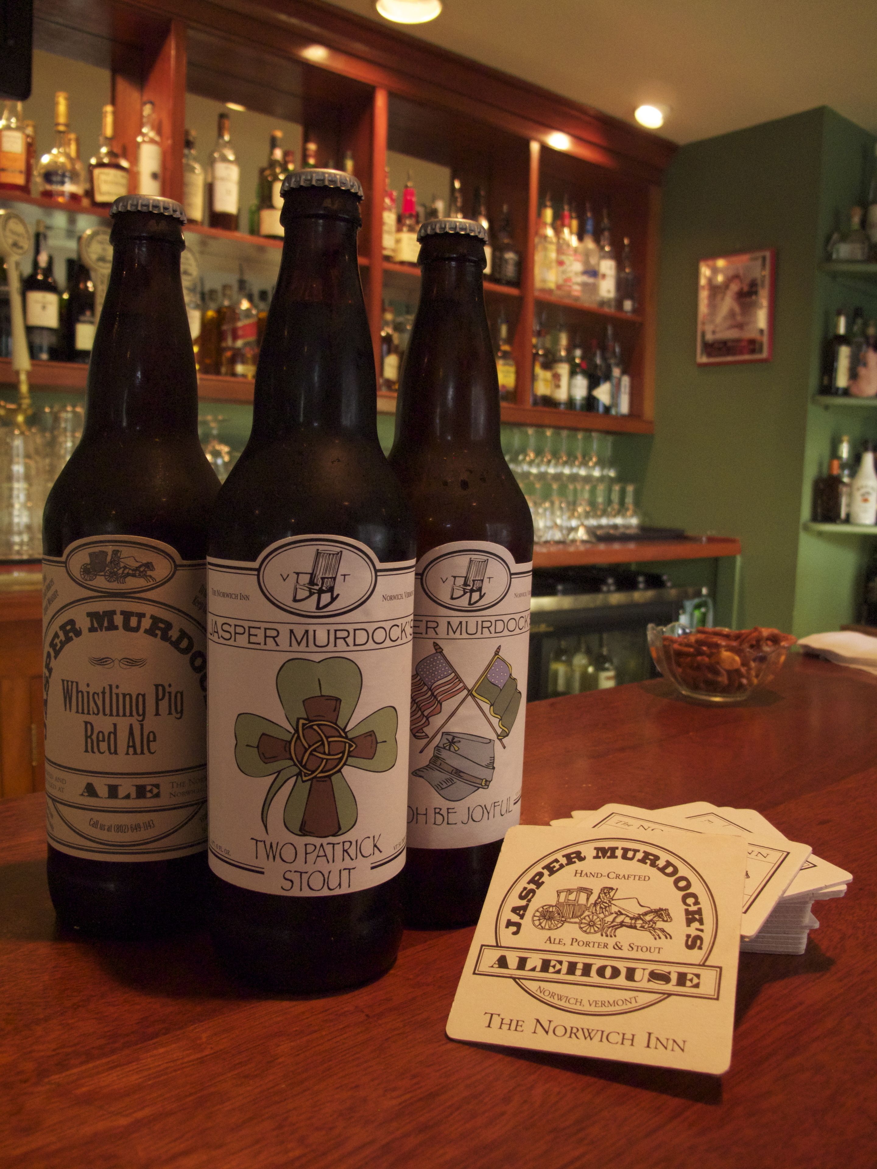 Three bottles of beer with labels, a stack of coasters, and a bar setting with shelves of bottles and glasses in the background.