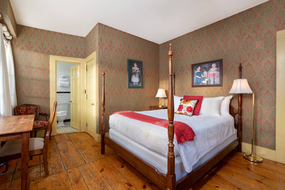 A bedroom with a four-poster bed, a wooden desk, a chair, and a floor lamp.