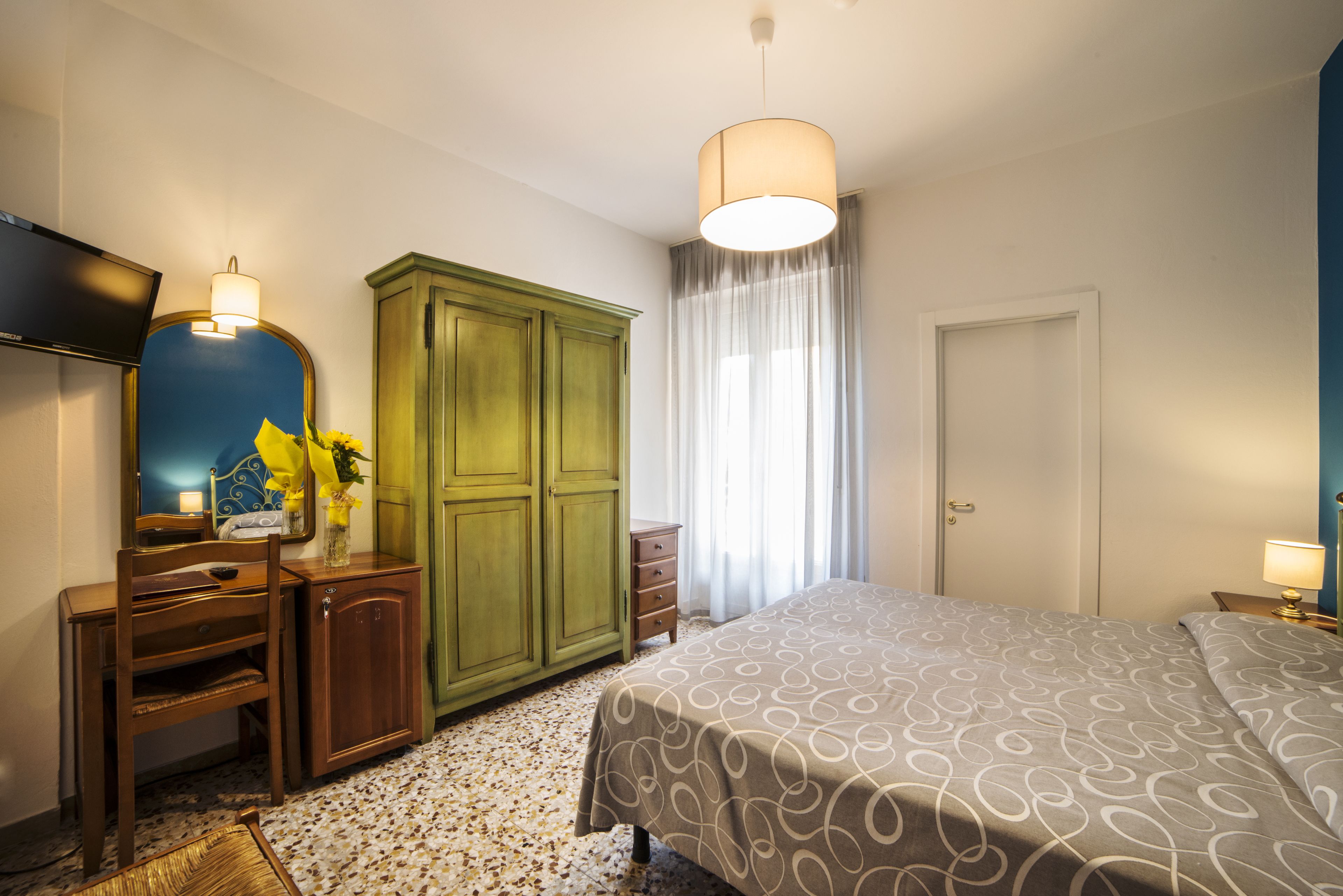 Une chambre à coucher comprenant un lit, un bureau avec une chaise, une armoire, un téléviseur et une fenêtre avec des rideaux.