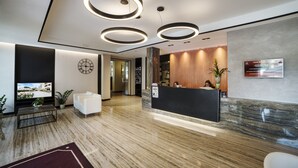 A modern hotel reception area with a marble floor, a reception desk, a clock, and a large screen displaying a scenic view. - Hotel Bisesti (Garda)