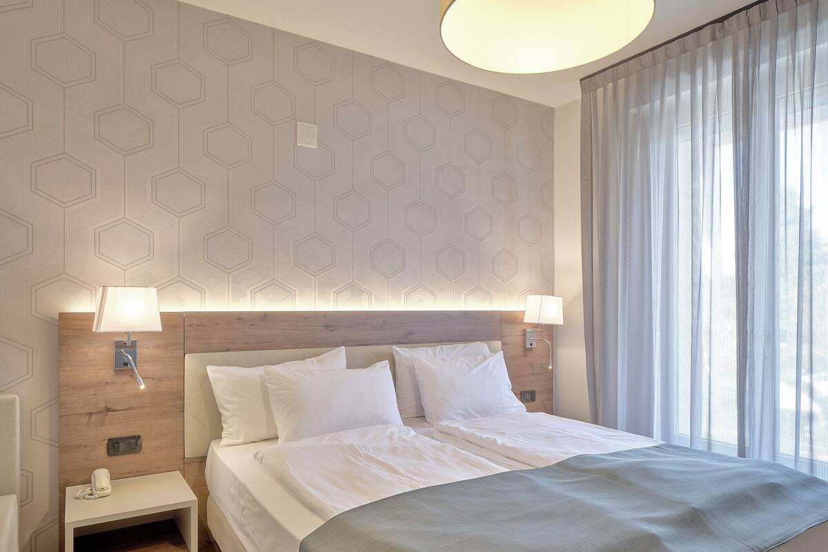 Une chambre d’hôtel avec un mur orné d’un motif géométrique, une tête de lit en bois, deux lampes de chevet, un lit avec du linge de maison blanc et une fenêtre avec des rideaux légers.