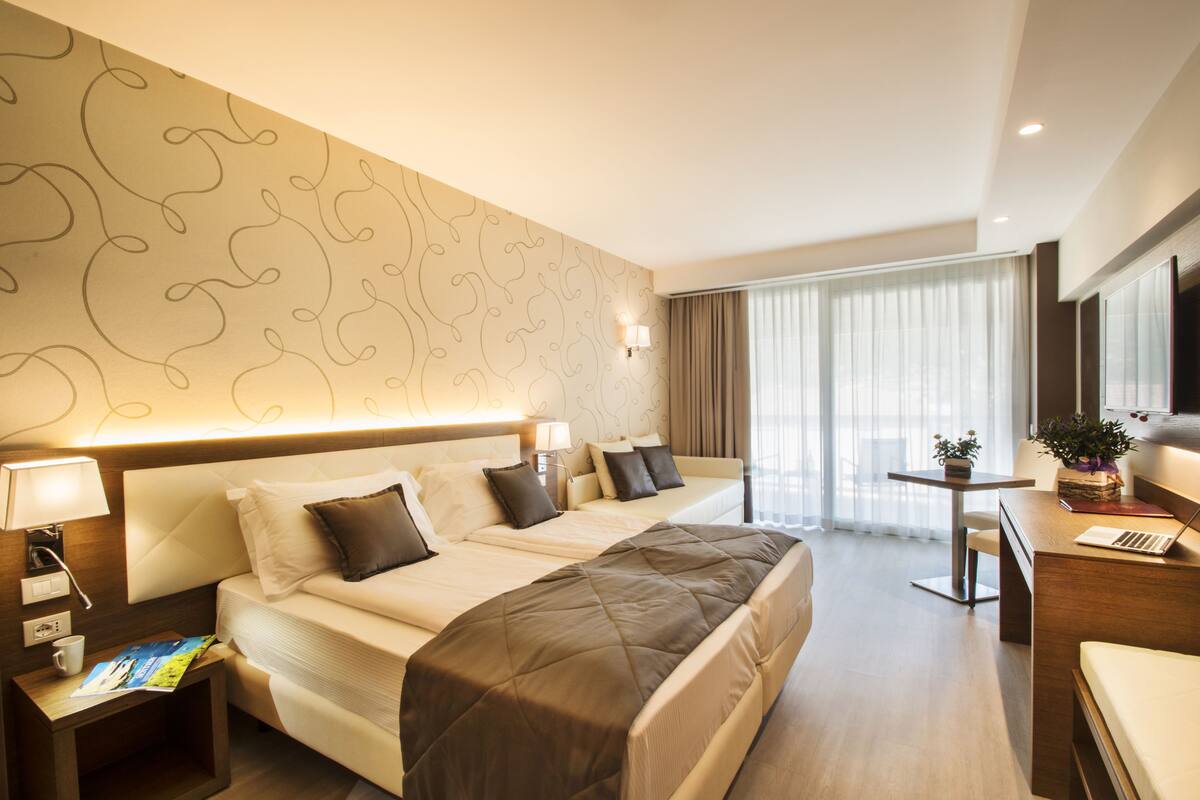Une chambre d’hôtel avec un grand lit, un bureau avec un ordinateur portable, une petite table avec une plante et une fenêtre avec des rideaux.