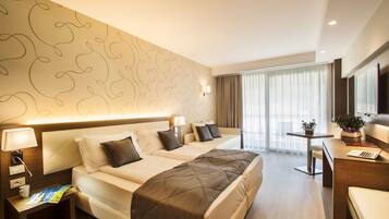 A hotel room with a large bed, a desk with a laptop, a small table with a plant, and a window with curtains.